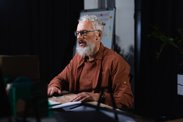 Fototapeta premium bearded businessman in eyeglasses holding smartphone while working in office at night