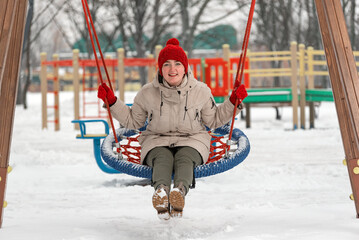 Young woman in warm clothes has fun in outdoors. Laughing girl rides swing on playground in winter