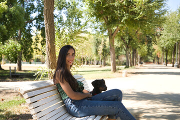 Obraz premium Young woman, brunette, slender, dressed in green t-shirt and jeans, sitting on a bench next to her dog, happy and in a loving attitude. Concept animals, pets, friends, love, dogs, breed.
