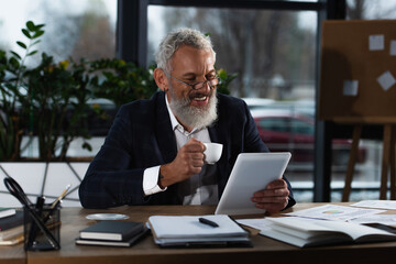 Obraz premium Cheerful mature businessman holding coffee cup while using digital tablet near documents in office