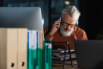 grey haired businessman in eyeglasses talking on smartphone near computers and blurred folders in...