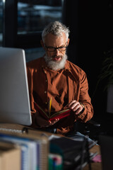 thoughtful bearded businessman looking in notebook near computer monitor on blurred foreground