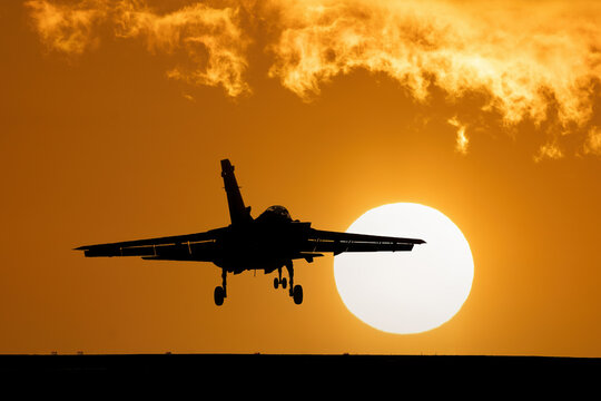 Fighter jet at sunset with orange sky and large setting sun after a combat mission. Flying over dramatic silhouette of fighter bomber jet aircraft