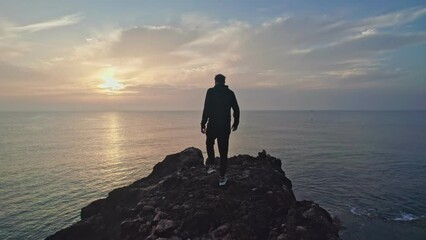 The camera follows a hipster millennial young man in a tracksuit running up a rock escarpment by the sea at sunset, throwing his hands up in the air, happy and drunk with life, youth and happiness