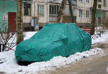 A car in the snow packed in a green case in the courtyard of the house, Iskrovsky Prospekt, St. Petersburg, Russia, December 2022