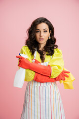 Obraz na płótnie Canvas stylish brunette housewife in red rubber gloves standing with rag and spray bottle in crossed arms isolated on pink