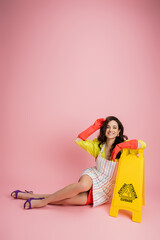 cheerful housewife in red rubber gloves looking at camera while sitting near wet floor sign on pink background