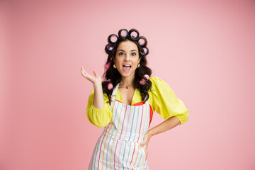 amazed housewife in striped apron and hair curlers standing with hand on hip isolated on pink