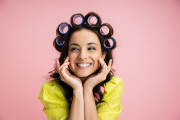 portrait of cheerful housewife in hair curlers holding hands near face while looking away isolated...