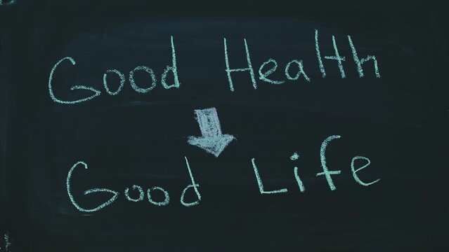 Child writing words good health and good life with arrow leading from one phrase to another with chalks on a blackboard in timelapse. Hand of a kid in pink clothes writing healthy lifestyle message on