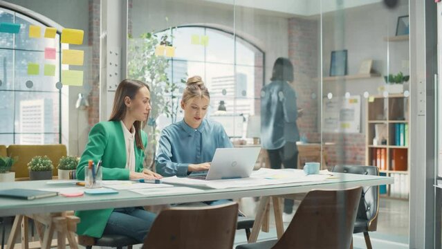 Portrait of Two Creative Colleagues Collaborating and Pointing at the Laptop Screen in Office Hub. White Female Designer Discussing Work with Assistant and Choosing Marketing Strategy