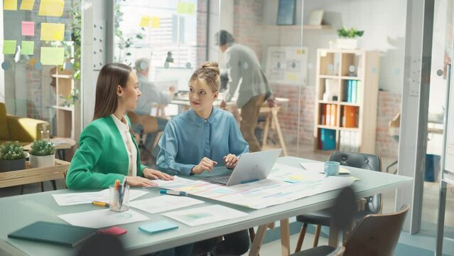 Two Creative Colleagues Discussing and Pointing at a Laptop in Modern Casual Office. Caucasian Sales Manager Discussing Project Plan with Female Legal Assistant and Taking Notes