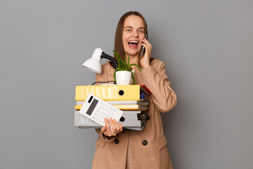 Portrait of excited amazed woman wearing beige jacket, holding paper folders posing isolated over...