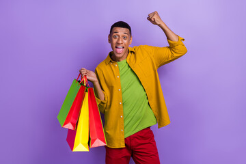 Photo of ecstatic good mood guy dressed yellow shirt holding shopping bags raise fist up shout yeah isolated on violet color background