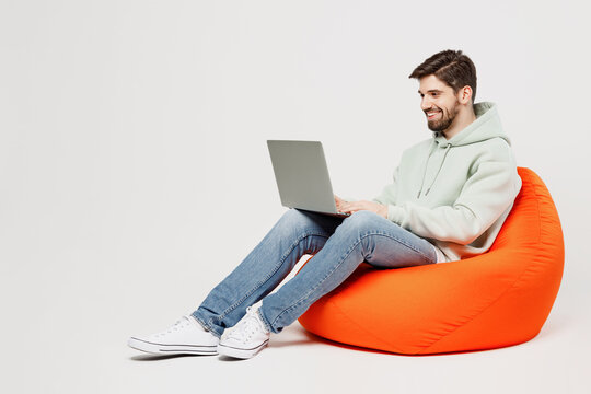 Full body young programmer happy caucasian IT man in mint hoody sit in bag chair hold use work on laptop pc computer isolated on plain solid white background studio portrait People lifestyle concept