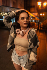 Beautiful woman with fashionable glasses and pink headphones in a fashion sweatshirt with a top and ripped jeans shorts walks in the night city with bokeh light