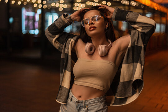 Fashionable beautiful hipster girl with fashion glasses and pink headphones in stylish clothes with a top and a plaid shirt walks on the street at night
