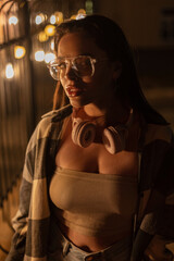 Stylish beautiful fashionable woman with eyewear glasses and headphones in a trendy top with a fashion plaid shirt walks in the night city with bokeh lights