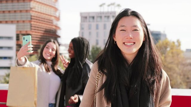 On foreground, one young asian brunette woman smiling looking at camera, with perfect white teeth, at background two friends taking a selfie photo after shopping sale day. Close up portrait of a happy