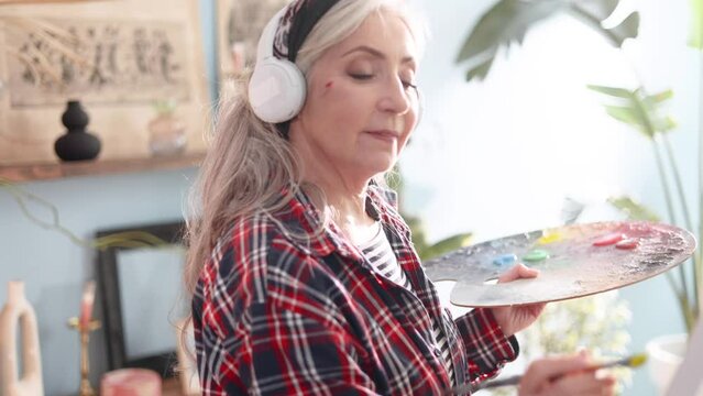 Senior woman artist listen music with headphones while painting picture on canvas creating artwork in sunny creative workshop Mature grey haired lady enjoy leisure time with art hobby