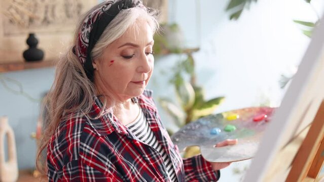 Portrait of charming senior woman artist painting picture on canvas creating artwork in the light sunny creative workshop Stylish mature grey haired lady enjoying leisure time with art hobby indoors