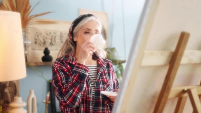 Dreamy soft portrait of charming stylish senior grey haired woman artist painter look at picture on canvas with cup of coffee or tea when finished artwork in the sunny creative workshop indoors