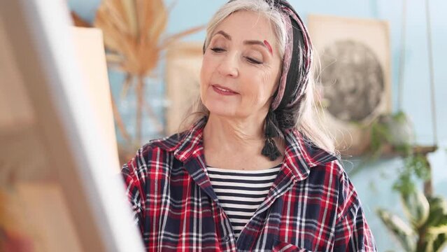 Portrait of charming senior woman artist painting picture on canvas creating artwork in the light sunny creative workshop Stylish mature grey haired lady enjoying leisure time with art hobby indoors