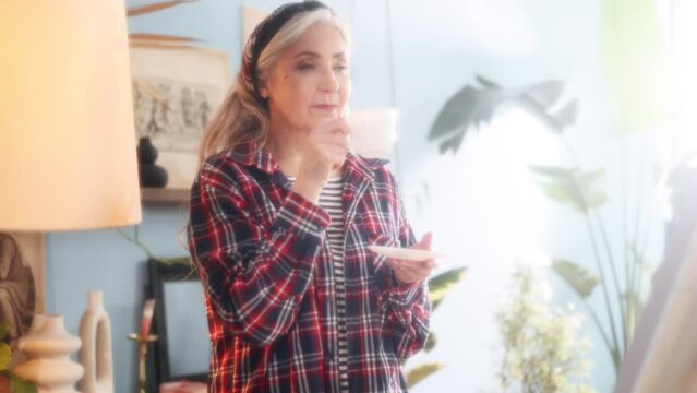 Dreamy soft portrait of charming stylish senior grey haired woman artist painter look at picture on canvas with cup of coffee or tea when finished artwork in the sunny creative workshop indoors