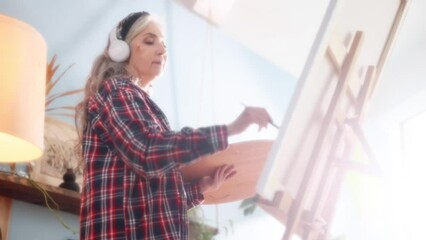 Dreamy soft of senior woman artist listen music with headphones while painting picture on canvas creating artwork in sunny creative workshop Mature grey haired lady enjoy leisure time with art hobby