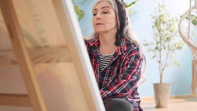 Charming senior woman artist painting picture on canvas creating artwork sits on floor in the light sunny creative workshop Stylish mature grey haired lady enjoying leisure time with art hobby indoors