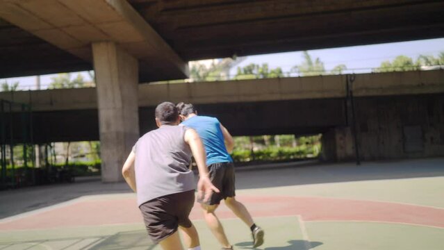 Two man athlete playing streetball match shooting and defense basketball on outdoors court together in sunny day. Sportsman do sport training basketball at street court under highway in the city.