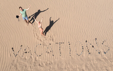 View from above to the dunes with word "vacations"on a sand. Summer holidays background. Man and woman posing near text"VACATIONS" in a sandy beach. VACATIONS concept