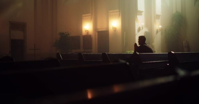 Wide Shot of Devout Christian Man Sits Piously In a Church, Folding Hands For Praying, Seeks Guidance From His Religious Faith and Spirituality. Spirit of Christianity and Hope for Repentance