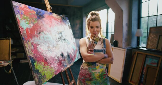 Confident modern woman artist looking at camera and smiling standing next to abstract impressionist artwork. Young artist in paint-stained apron stands in studio. Portrait of artist with arms crossed