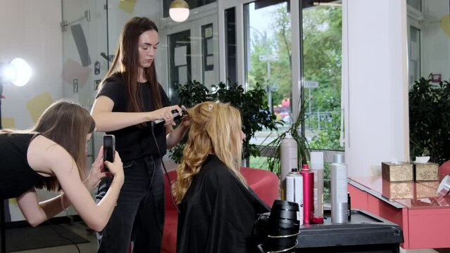 Young stylist does styling curls for blonde client using modern curling iron in beauty salon. Receptionist takes photos with phone of hairdresser working process