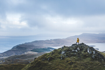 A person in a yellow jacket standing on top of a mountain overseeing the landscape of Isle of Skye...