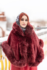 A Slavic woman in a national colored scarf, a fur coat and warm mittens on the porch of a wooden house. Winter, snowfall