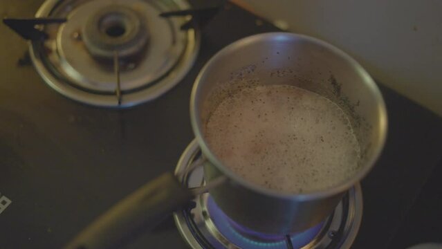 Close-up of man hand preparing  coffee in steel utensils on black gas stove in kitchen. male shakes boiling on heat temperature tea vessel by holding pan handle