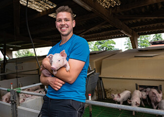 Junger Landwirt  steht vor einer Ferkelbucht mit Ferkeln und trägt ein Ferkel auf den Arm.