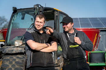 Vater und Sohn, beide Landwirte, stehen am Trecker der einen Futtermittelwagen angehängt hat, sehen zufrieden aus.