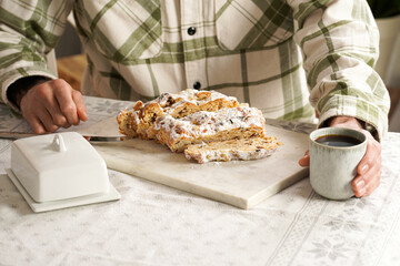 Man in a checkered shirt sitting at the table ready to eat traditional german christmas pastry christstollen on a marble cutting board, coffee mug, butter plate on the table cloth