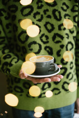 Young stylish person in a green sweater with leopard print on it, holding a cup of freshly brewed cappuccino coffee with a beautiful latte art on it, with festive fireflies on the background