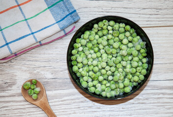 Fresh, frozen green peas in a black bowl, wooden spoon, and cloth on a white table.