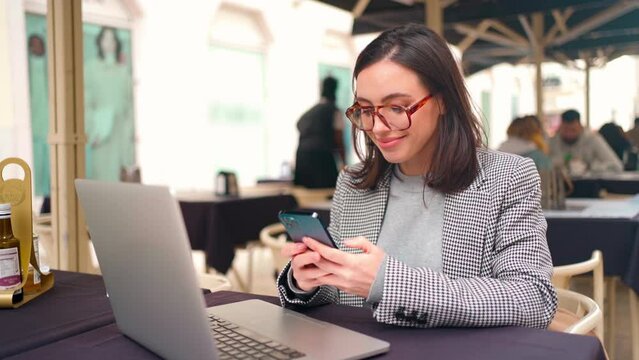 Woman working with laptop use smartphone sitting Street Cafe in european city side view. Caucasian female business woman freelancer using laptop for remote work while sitting table restaurant