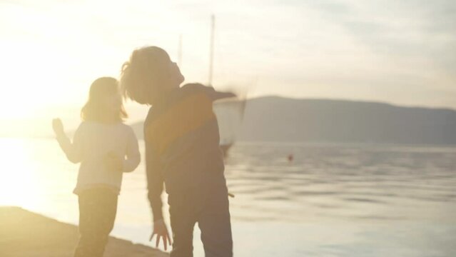 Boy and girl on a pier by the sea throws stones into the water against the backdrop of sunset