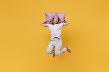 Fototapeta na wymiar Full body young woman she wears purple pyjamas jam sleep eye mask rest relax at home jump high hold in hand pillow behind neck isolated on plain yellow background studio portrait. Night nap concept.