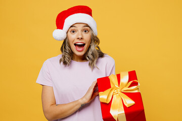 Young surprised shocked woman she wears purple pyjamas jam Santa hat rest relax at night home nap hold red present box with gift ribbon bow isolated on plain yellow background. New Year 2023 concept.