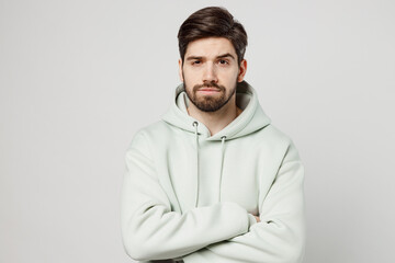 Young displeased sad caucasian man wear mint hoody look camera holding hands crossed folded waiting for apologises isolated on plain solid white background studio portrait. People lifestyle concept.