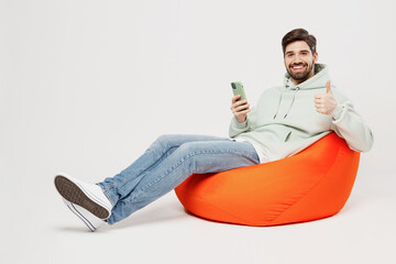 Full body young satisfied happy man wear mint hoody sit in bag chair hold in hand use mobile cell...