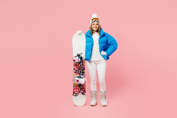 Full body snowboarder woman in blue suit goggles mask hat ski padded jacket hold snowboard stand akimbo isolated on plain pastel pink background Winter extreme sport hobby weekend trip relax concept